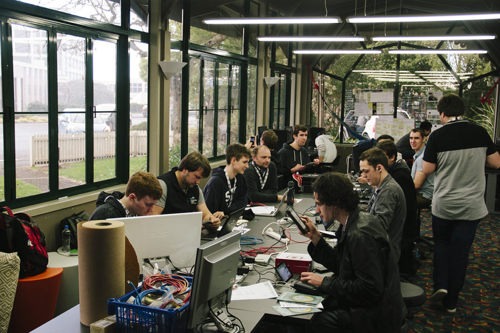 A group of young men, all sitting at a long desk, working on various things at computers and laptops